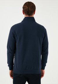 Giacca pullover blu navy con colletto alto, maniche lunghe, orlo e polsini a coste; realizzata in un tessuto morbido e strutturato.