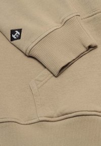 Beige Kapuzenpullover mit gerippten Bündchen und Saum. Verfügt über eine Fronttasche und ein schwarzes Logo-Patch am Ärmel. Weicher, strukturierter Stoff.