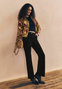 Jachetă tricotată cu model floral în galben și roșu peste un top scurt negru, asortată cu pantaloni din catifea închiși la culoare și cizme cu tocuri negre.