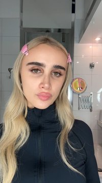 Giovane donna con lunghi capelli biondi fissati da mollette rosa, che indossa un top nero con zip, in piedi in un bagno con piastrelle bianche.