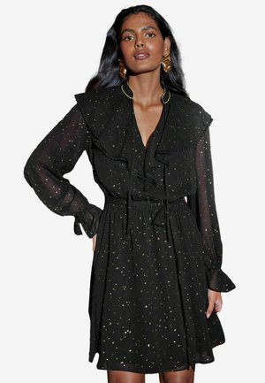 Robe noire avec un col à volants, des manches longues en tissu transparent et une taille cintrée. Elle présente des éclats dorés et une jupe fluide.