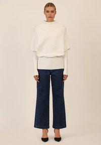 Weißer gerippter Rollkragenpullover mit bauschigen Ärmeln, kombiniert mit dunkelblauen, weit geschnittenen Jeans mit subtilen Verzierungen. Schwarze Absätze vervollständigen das Outfit.