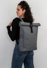 Rucsac gri cu capac rulat, fabricat dintr-un material texturat, având o cataramă neagră, curele ajustabile și buzunare laterale pentru depozitare.