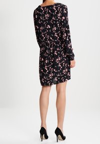 Robe noire à manches longues avec un motif floral rose, texture douce, longueur au-dessus du genou et taille cintrée, associée à des talons hauts noirs.