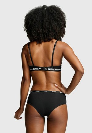 Frau mit lockigem Haar, die schwarzen Puma-BH und Slips trägt, von hinten vor hellem, einfarbigem Hintergrund gezeigt.