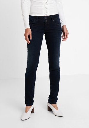 Person iført mørke slim-fit jeans, hvid langærmet top med knapper og hvide sko med blokhæle, stående på et hvidt gulv.
