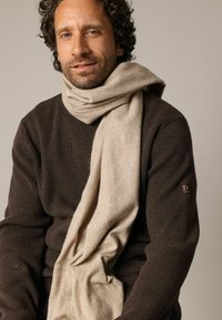 Écharpe en laine beige enroulée autour du cou d'un homme, complétant un pull à côtes marron foncé. Texture notable et tombé doux.