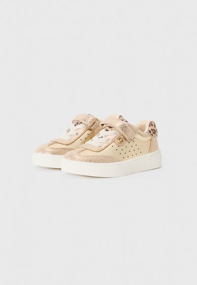 Zapatillas beige con acentos brillantes, textura suave, diseño perforado y correa ajustable de velcro. Suela blanca con una marca sutil.