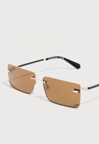 Occhiali da sole rettangolari senza montatura con lenti marroni, sottile ponte dorato e aste nere, appoggiati su una superficie bianca.