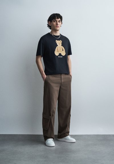 Junger Mann, der ein schwarzes T-Shirt mit einem Teddybär-Motiv, braune Cargohosen, weiße Turnschuhe und eine silberne Kettenhalskette trägt, steht vor einer einfachen Wand.