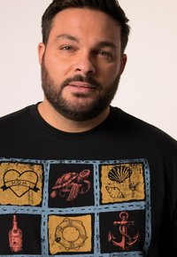 T-shirt noir avec un design graphique coloré composé de carrés représentant un cœur, une tortue, une coquille, une bouteille de rhum, une bouée de sauvetage et une ancre.