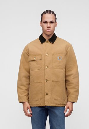Jonge man met gevlochten haar, gekleed in een beige jas met knopen, zwarte kraag en vier voorzakken, staand tegen een effen lichte achtergrond.