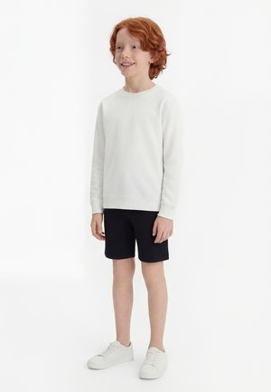 Garçon aux cheveux roux souriant, portant un sweat-shirt blanc, un short noir et des baskets blanches, debout devant un fond blanc uni.