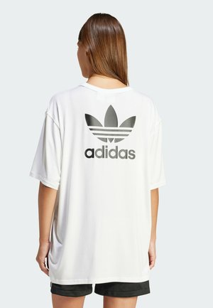 Camiseta blanca de gran tamaño hecha de algodón, con un gran logo negro de Adidas y un diseño de tres rayas en la parte posterior, con mangas cortas.