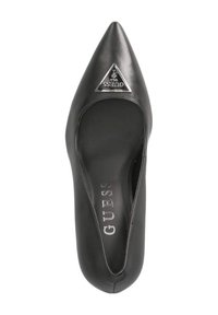 Pump nera in pelle sintetica con punta affusolata, caratterizzata da un accento logo triangolare in argento e una texture liscia. All'interno si trova il marchio "GUESS".