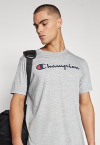 Graues Baumwoll-T-Shirt mit kurzen Ärmeln, das ein blaues und rotes "Champion"-Logo auf der Brust präsentiert. Schwarzer Riemen einer Tasche über der Schulter.