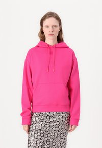 Felpa cappuccio rosa con tasca a canguro, lacci e logo tono su tono sul petto. Indossata con pantaloni a righe bianchi e neri.