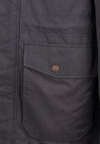 Vadderad svart jacka med en stor lappficka med en metallknapp. Slät tyg med en subtil glans och minimalistisk design.