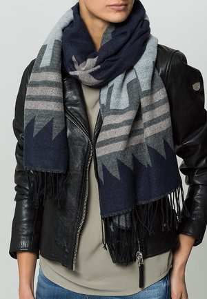 Femme portant une veste en cuir noire, un t-shirt gris, un jean bleu et une grande écharpe à motifs bleu marine, gris et beige avec des franges.