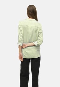 Camisa de botón de rayas verdes y blancas con mangas largas, ajuste relajado, detalle en el yugo trasero y cintura curva. Llevada con pantalones negros.