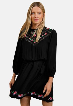 Kvinde med langt blond hår iført en sort kjole med pink blomsterbroderi på bryst og kant, stående mod en ensfarvet baggrund.