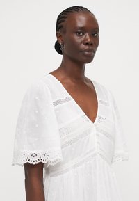 Hvit bomullsdrakt med V-hals, korte pufferm, og broderte detaljer. Har dekorative knapper og et teksturert stoff med utskjæringer.