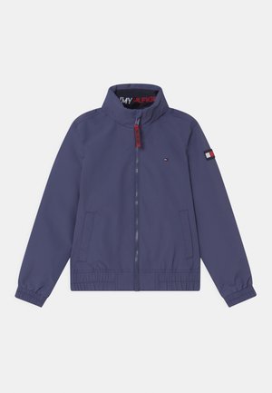 Mørkeblå, let jakke med høj krave, lynlås foran, elastiske manchetter og en logo-patch på venstre ærme. Glat tekstur.