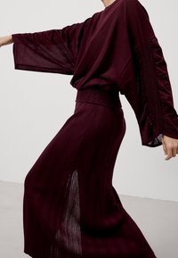 Viininpunainen neulottu yläosa leveillä hihoilla ja resorilla vyötäröllä, yhdistetty samanlaiseen laskostettuun hameeseen, jossa sivuhalkio. Tasainen ja sileä koostumus kauttaaltaan.