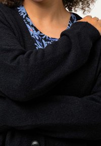 Cárdigan de punto negro con una textura suave, que presenta un detalle de botón. Debajo, se puede ver parcialmente una blusa con un estampado floral azul.