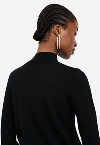 Maglione nero a maglia con colletto, dettagli di cucitura sottili e una silhouette aderente. Orecchini in cristallo grandi aggiungono un accento di contrasto.
