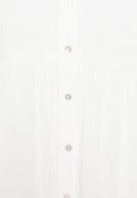 Gros plan d’un vêtement blanc avec une rangée verticale de boutons translucides au centre.