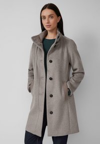 Vrouw in een middelgrote beige wollen jas met zwarte knopen, staand met haar handen in de zakken tegen een effen grijze achtergrond.
