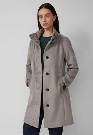 Femme portant un manteau en laine beige mi-long avec des boutons noirs, debout les mains dans les poches contre un fond gris uni.