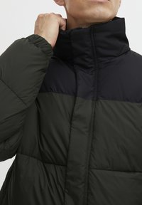 Pufjakas ar melnu augšējo daļu un tumši zaļu apakšējo daļu, ar augstu apkakli, quiltēta struktūra un elastīgām aprocēm.