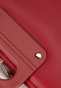 Czerwona skórzana torba o teksturowanym wykończeniu, z akcentem w złotym kolorze i zamknięciem na zamek. Gładki uchwyt o strukturalnym designie.