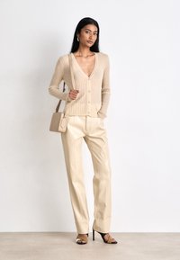 Cardigan beige a coste con scollatura a V profonda, frontale con bottoni, abbinato a pantaloni in pelle lucida beige e décolleté con cinturini bicolori. Piccola borsa a tracolla.