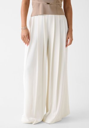 Kobieta ubrana w białe spodnie z wysokim stanem o szerokich nogawkach z przednimi zakładkami oraz beżową bluzkę, stojąca na jednolitym białym tle.