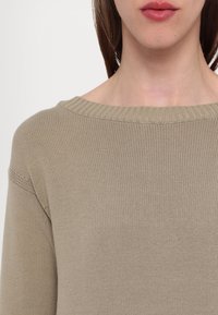 Beige strikkegenser med bred ribbestrikket hals, teksturert mønster, lange ermer og løs passform.