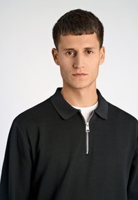 Polo shirt nero a metà zip con superficie testurizzata, caratterizzato da un colletto piatto e dettagli in cerniera argentata sullo scollo.