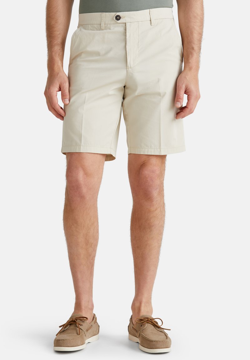 Beige men's knee-length shorts with button and zipper closure, worn with green shirt and brown lace-up boat shoes.