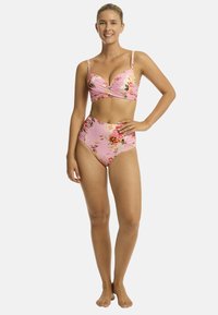 Modelka w różowym bikini w kwiaty, uśmiechnięta i pewna siebie, pozująca z jedną ręką na biodrze na neutralnym tle.