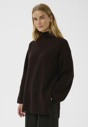 Dark brown knitted turtleneck sweater with a loose fit, ribbed texture, and side slits, paired with black pants.