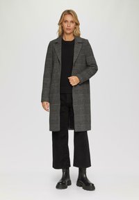 Grau karierten langen Mantel mit Schlitzkragen, Knopfverschluss und seitlichen Taschen, kombiniert mit einem schwarzen Pullover und schwarzen Hosen. Schwarze Stiefeletten.
