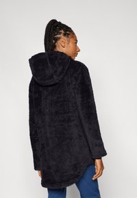 Schwarzer plüschiger Fleece-Mantel mit Kapuze, der eine weiche Textur und einen längeren Schnitt aufweist, von hinten betrachtet. Einfaches Design ohne sichtbare Hardware.