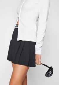 Vit långärmad golfpullover med strukturerad design, kombinerad med en svart plisserad golfsärk. Håller en svart golfklubba.