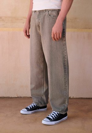 Person iført løstsiddende grå jeans og sort-hvide sneakers, stående op ad en beige væg med armene afslappet ned langs siden.