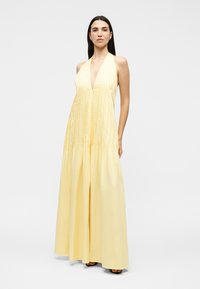 Femme portant une robe longue sans manches, jaune pâle, avec un décolleté en V profond et des détails plissés, debout devant un fond uni.