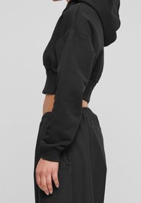 Svart kort hoodie i mjukt tyg, med huva och vida ärmar, matchad med lösa svarta byxor, visad från sidan.