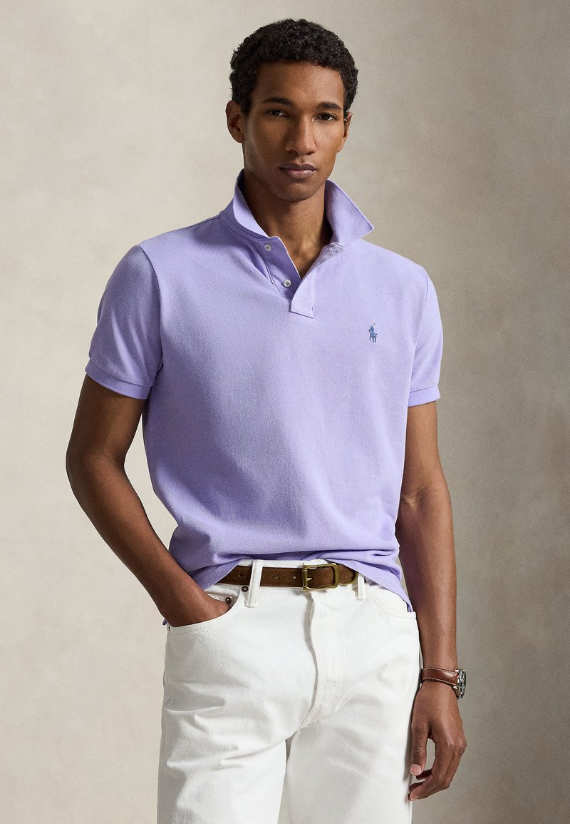 Joven con una camisa tipo polo color lila claro con cuello levantado, pantalones blancos, cinturón marrón y reloj de pulsera, posando frente a un fondo neutro.