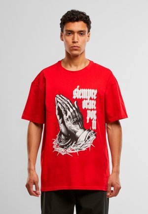 Nuori mies, jolla on päällään punainen t-paita, jossa on mustat rukoilevat kädet ja espanjankielinen teksti "siempre orar por ti", jota ympäröi piikkikruunu.